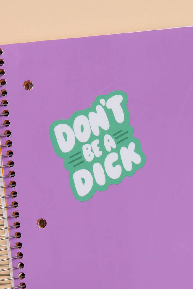 Purple notebook with 'Don't be a dick' sticker on a beige background with stationery items.