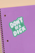 Purple notebook with 'Don't be a dick' sticker on a beige background with stationery items.