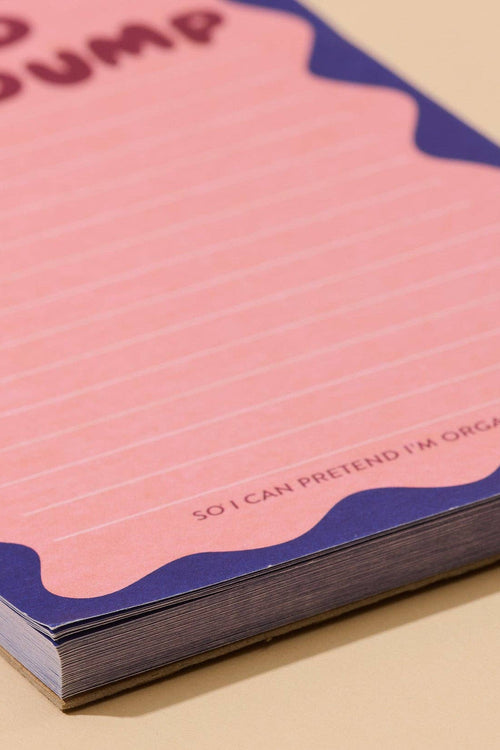 A pink lined notepad with a wavy dark blue border, shown at an angle on a light beige background. The top reads 'ADHD BRAIN DUMP' and the bottom edge includes the phrase 'SO I CAN PRETEND I'M ORGANIZED'.