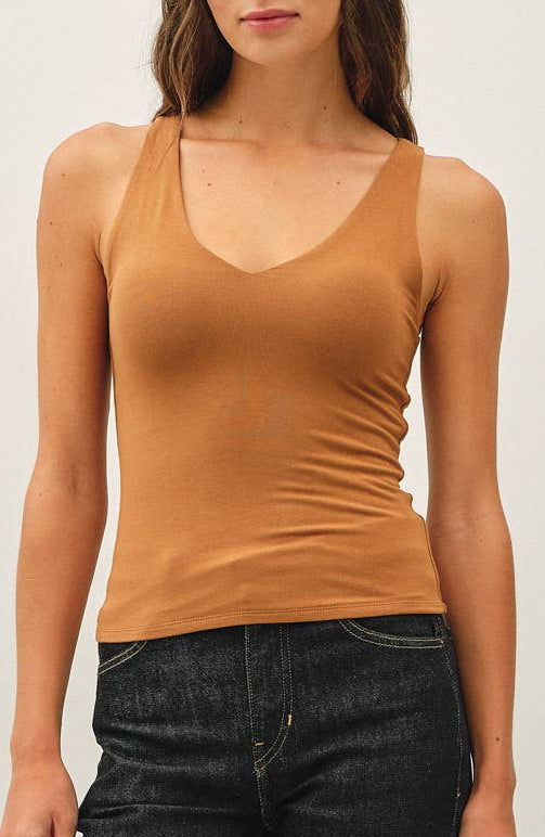 A woman modeling a fitted tan sleeveless tank top with dark wash jeans and dark sneakers, standing against a plain off-white studio background.