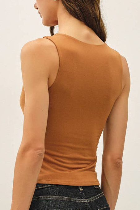 A woman shown from the back wearing a fitted sleeveless brown top, dark blue jeans, and dark slip-on shoes, standing against a plain off-white studio background.