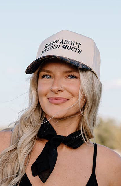Woman wearing a black tank top, denim shorts, and a white cap with text outdoors.