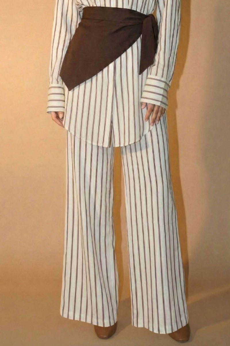 Woman wearing a striped shirt and wide-leg pants with a brown belt against a beige background