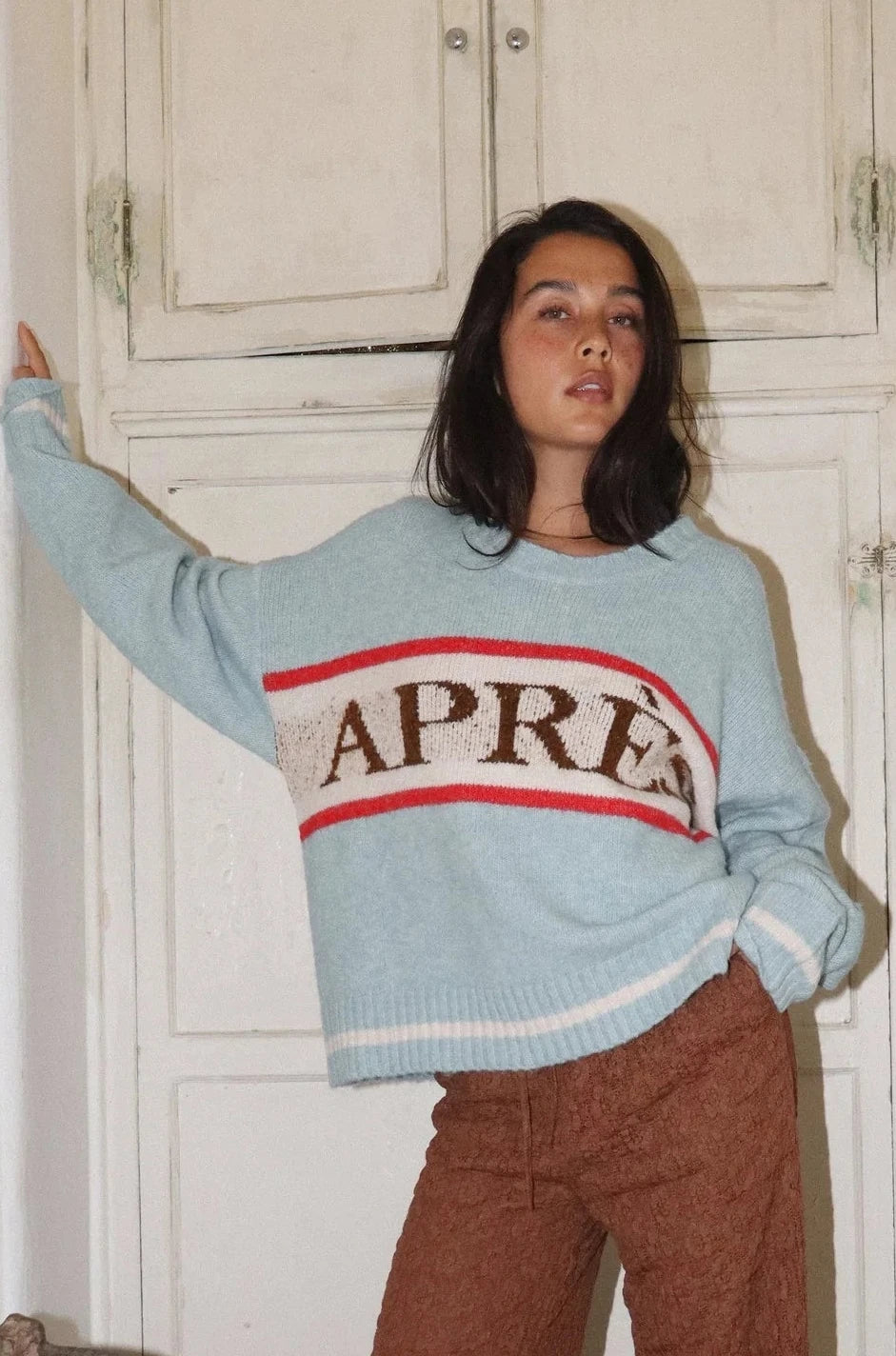 Person wearing a light blue sweater with 'APRÈS' text and brown pants in an indoor setting.