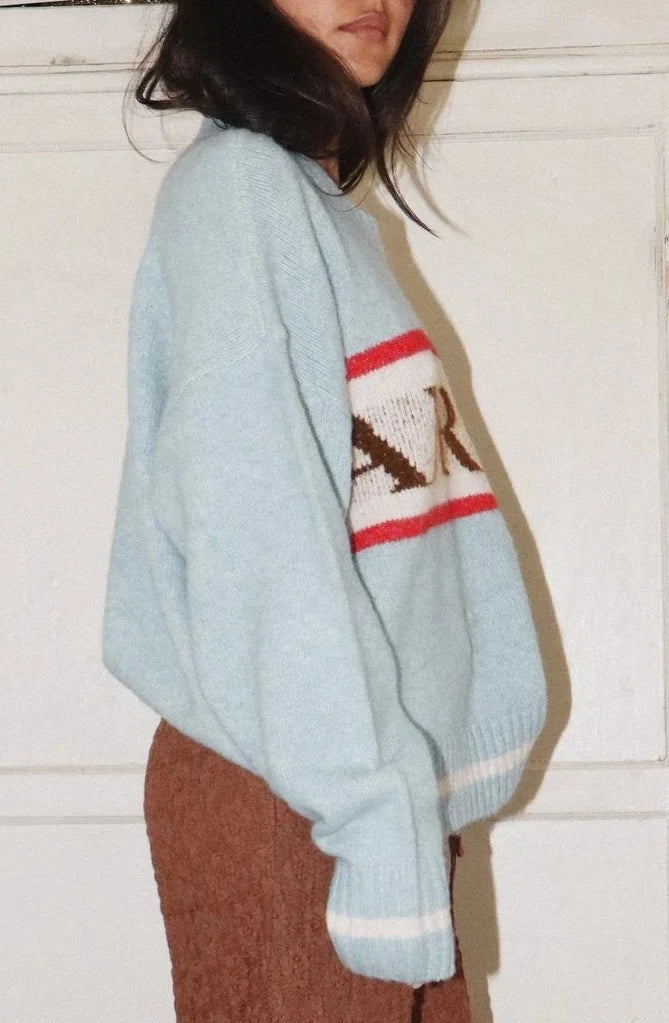Person wearing a light blue sweater with a graphic design, standing in a room with wooden furniture.