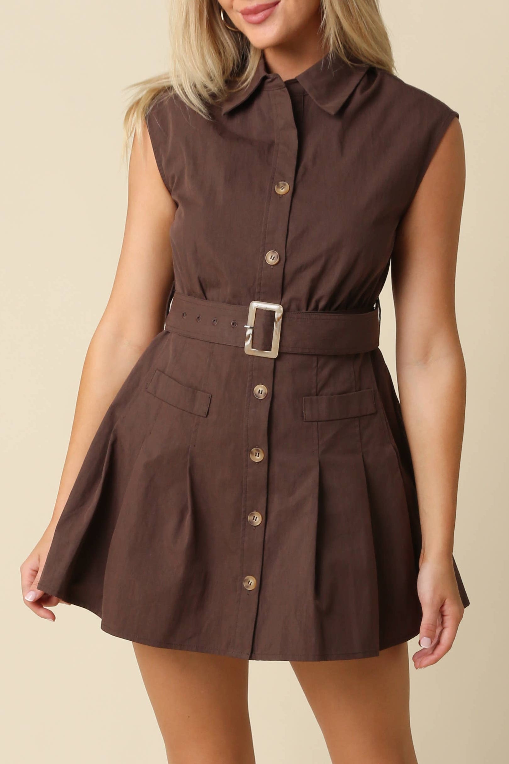 A woman modeling a sleeveless brown mini dress with a pointed collar, button front, and matching belt with a square buckle, photographed against a plain beige studio background.