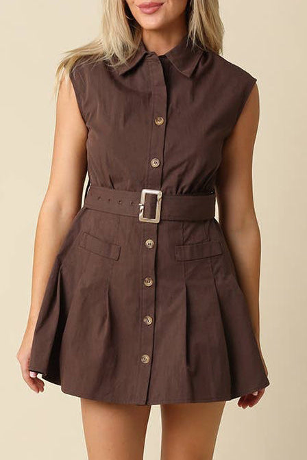 A woman modeling a sleeveless brown mini dress with a pointed collar, button front, and matching belt with a square buckle, photographed against a plain beige studio background.