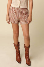 Woman wearing a beige sleeveless button-up top, plaid shorts, and brown boots on a beige background