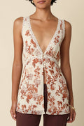 A woman modeling a sleeveless cream camisole top with a rust-brown floral print and lace trim, paired with dark brown pants, against a solid beige background.