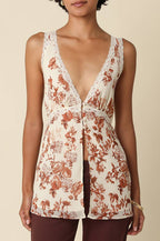 A woman modeling a sleeveless cream camisole top with a rust-brown floral print and lace trim, paired with dark brown pants, against a solid beige background.