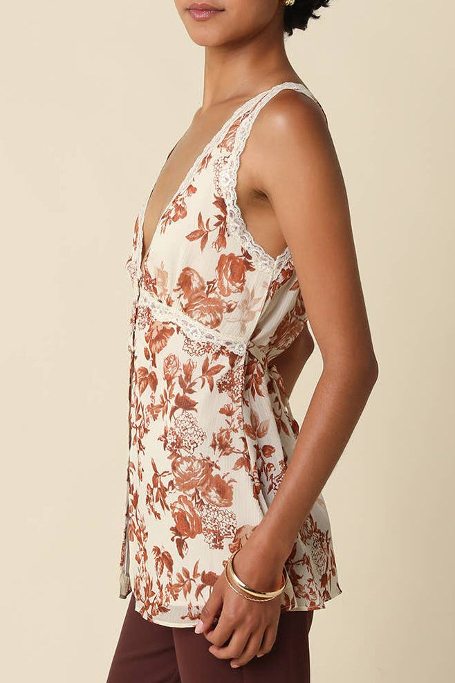 A woman wearing a sleeveless cream top with a rust-brown floral toile pattern and dark brown pants, photographed against a solid light beige studio background.