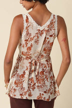 A woman shown from the back wearing a sleeveless cream-colored top with a rust-orange floral pattern and lace trim, paired with dark brown pants, against a solid beige background.