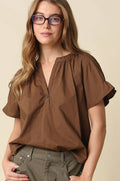 A woman with glasses wearing a brown short-sleeve blouse with a gathered neckline and olive green pants, standing against a solid beige background.