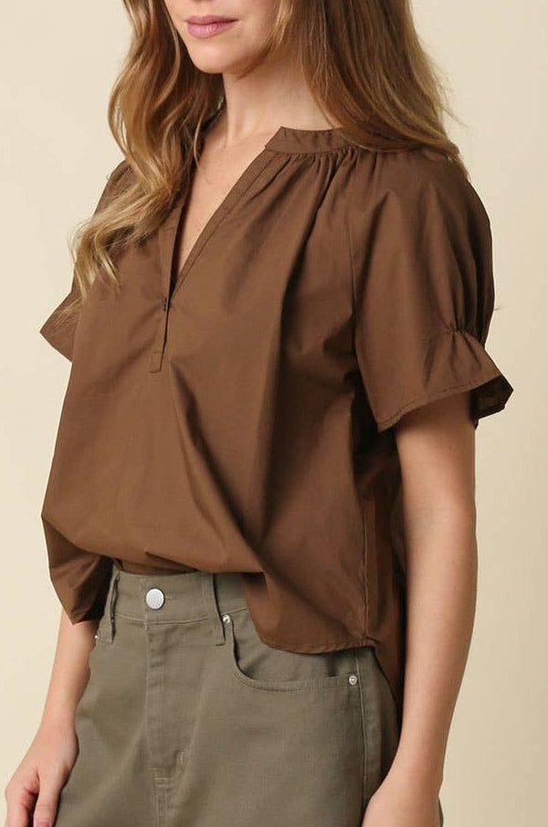 A woman with long light brown hair and purple-framed glasses wearing a brown short-sleeve blouse with a gathered V-neckline, paired with olive green pants, against a solid beige background.