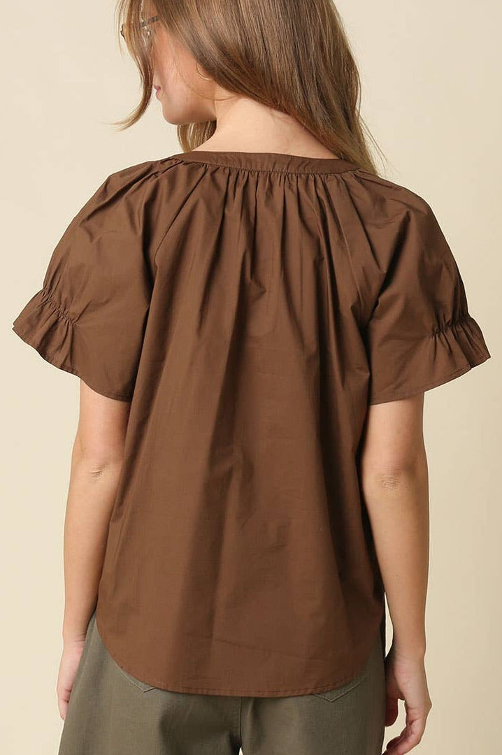 A woman shown from the back wearing a brown short-sleeve blouse with a gathered, ruffled neckline and flared sleeves, paired with olive green pants, against a plain beige background.