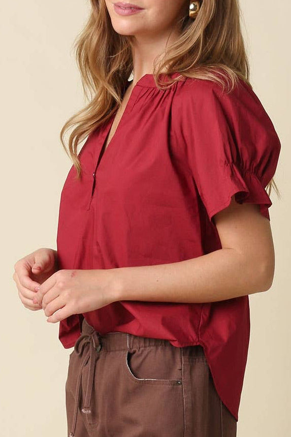 A woman modeling a burgundy short-sleeve blouse with puff sleeves and a gathered neckline, paired with brown pants, against a plain beige studio background.