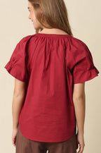 A woman shown from the back wearing a red short-sleeve blouse with a gathered, ruffled neckline and brown pants, set against a plain beige studio background.
