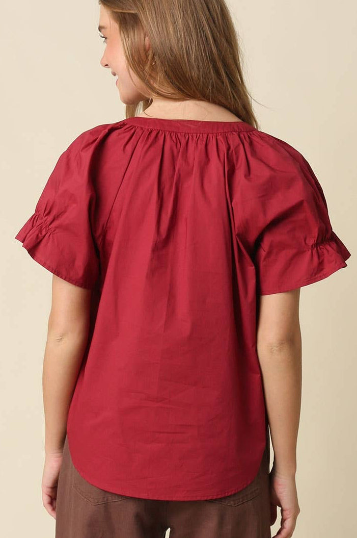 A woman shown from the back wearing a red short-sleeve blouse with a gathered, ruffled neckline and brown pants, set against a plain beige studio background.
