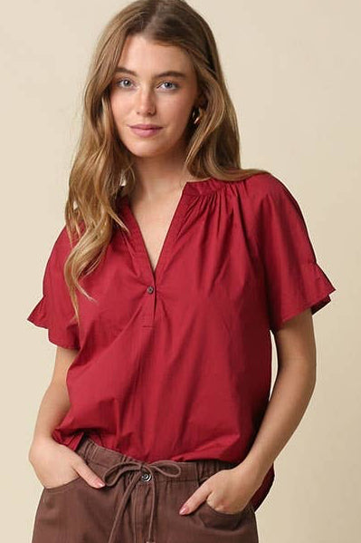 A woman modeling a red short-sleeve blouse with a gathered neckline and brown wide-leg pants, standing against a plain beige studio background.