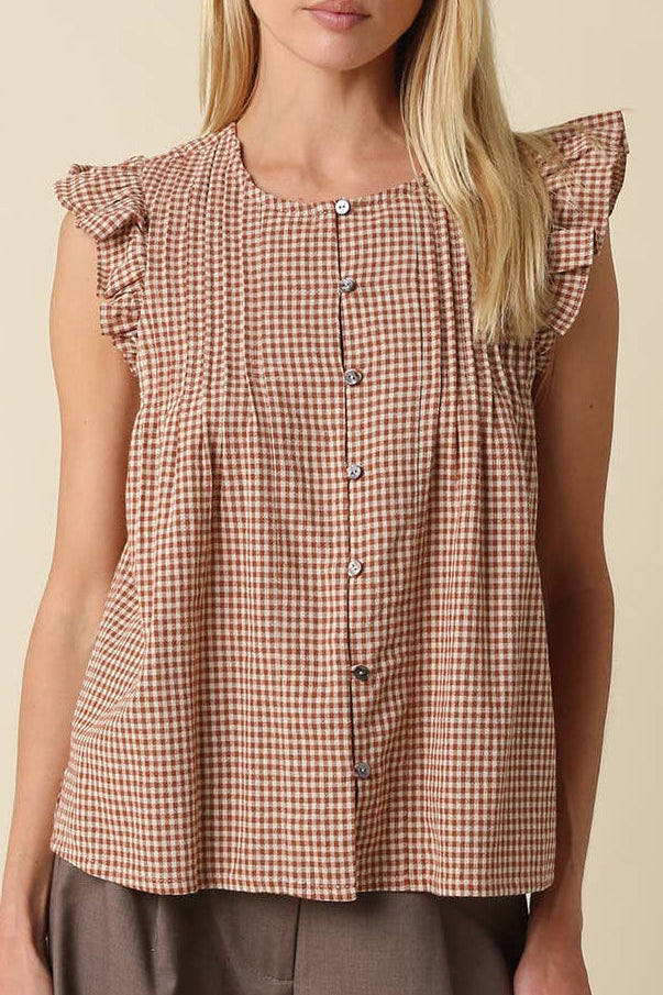 A woman modeling a sleeveless brown and beige gingham top with ruffled shoulder details and a button-down front, paired with brown pants against a solid beige background.