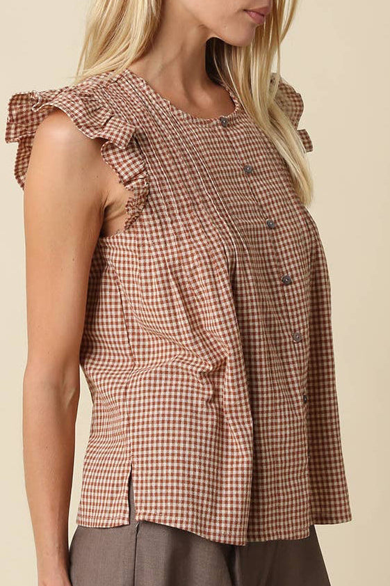 A woman wearing a brown and beige gingham sleeveless top with ruffled cap sleeves and a buttoned front, paired with brown pants, photographed against a solid beige background.