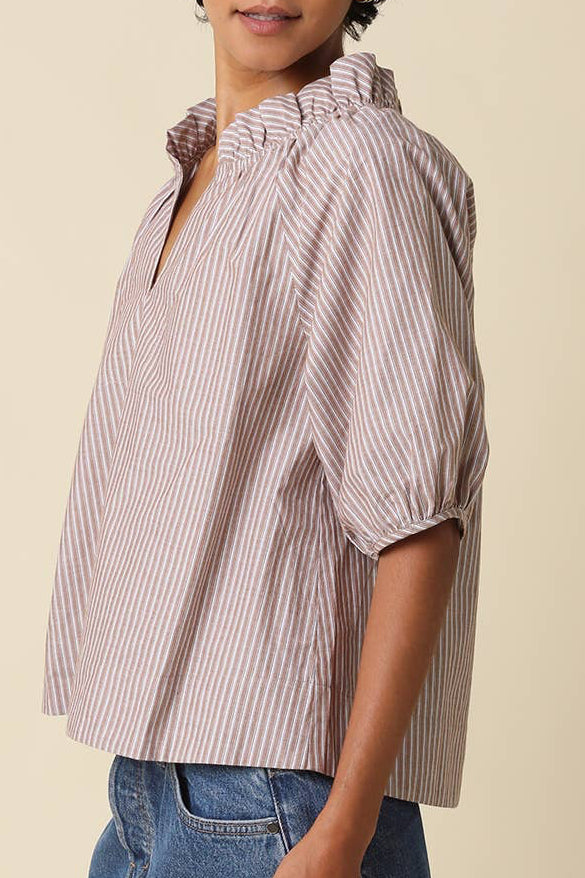 A woman modeling a short-sleeve button-up blouse with vertical pink and white stripes, paired with denim shorts, against a solid beige background.