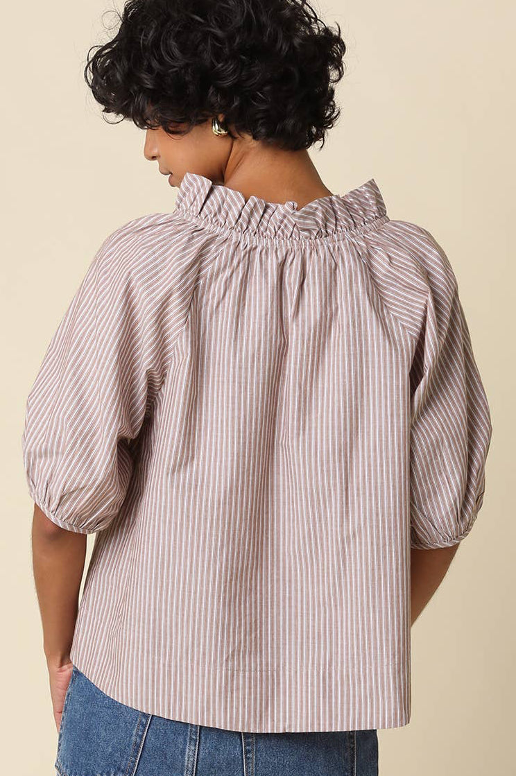 A woman shown from the back wearing a light pink and white vertically striped blouse with a smocked ruffled neckline and puff sleeves, paired with a blue denim skirt, against a solid beige background.