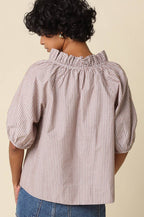 A woman shown from the back wearing a light pink and white vertically striped blouse with a smocked ruffled neckline and puff sleeves, paired with a blue denim skirt, against a solid beige background.