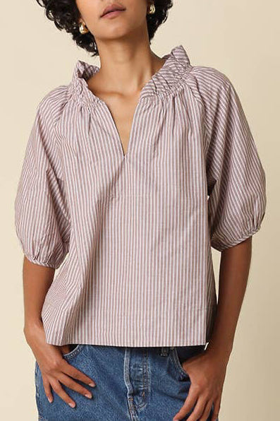 A model wearing a short-sleeved, puffed blouse with vertical pink and white stripes, paired with a denim mini skirt, white socks, and black loafers, standing against a solid beige background.