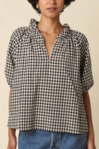 A woman wearing a black and white gingham check short-sleeve blouse with a ruffled split neckline, paired with a blue denim skirt, against a solid beige background.