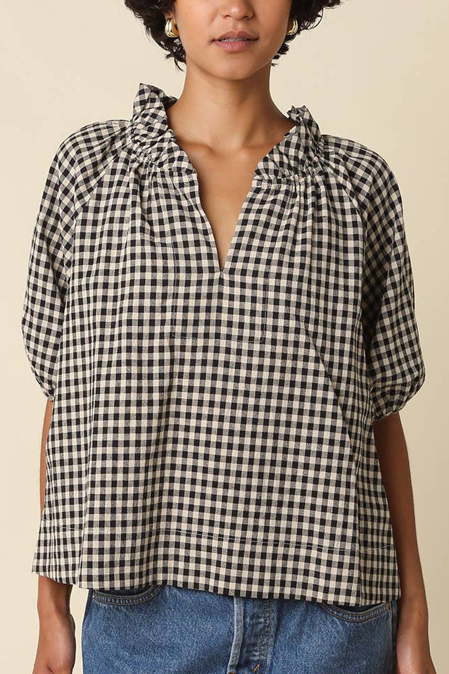 A woman wearing a black and white gingham check short-sleeve blouse with a ruffled split neckline, paired with a blue denim skirt, against a solid beige background.