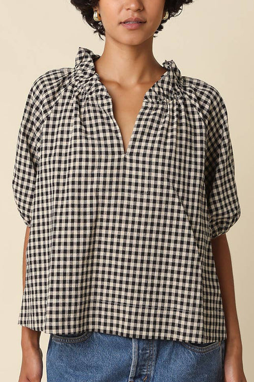 A woman wearing a black and white gingham check short-sleeve blouse with a ruffled split neckline, paired with a blue denim skirt, against a solid beige background.