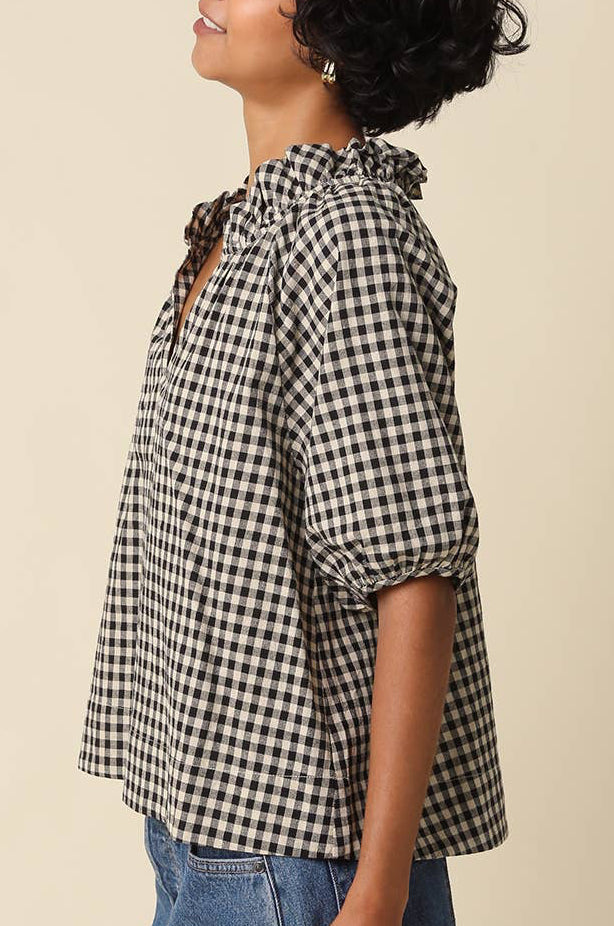 A woman wearing a black and white gingham tie-neck blouse with puff sleeves, paired with denim shorts, shown in profile against a solid beige background.