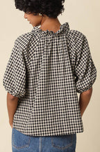 A woman shown from the back wearing a black and white gingham check blouse with puff sleeves and a ruffled neckline, paired with a denim skirt, against a plain beige background.