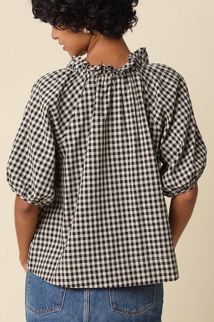 A woman shown from the back wearing a black and white gingham check blouse with puff sleeves and a ruffled neckline, paired with a denim skirt, against a plain beige background.