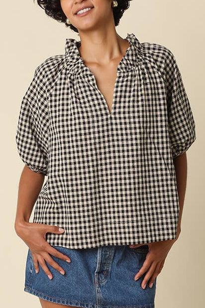 A woman modeling a black and white gingham short-sleeve button-up shirt with a ruffled collar, paired with a blue denim mini skirt, brown socks, and black loafers, against a solid beige studio background.