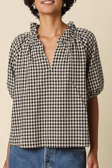A woman modeling a short-sleeve black and white gingham blouse with a split neckline, paired with a blue denim mini skirt, brown socks, and black loafers, against a solid beige background.