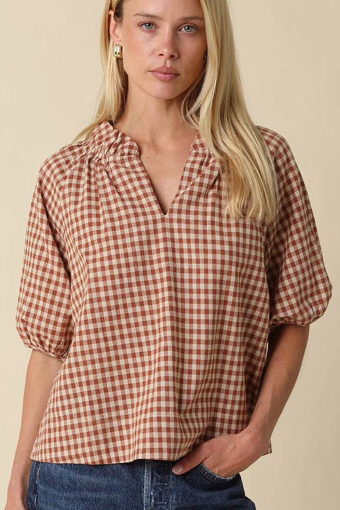 A woman modeling a short-sleeve blouse with a brown and beige gingham pattern, paired with blue jeans, against a solid beige background.