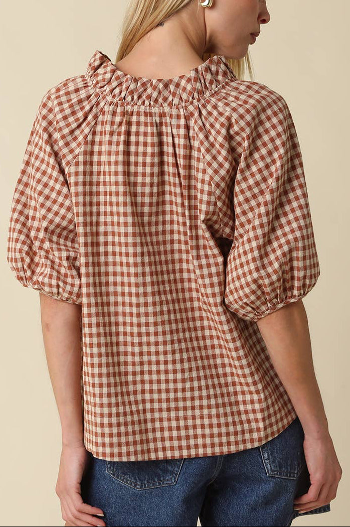 A woman shown from the back wearing a brown and beige gingham check blouse with puff sleeves and a ruffled neckline, paired with blue jeans against a solid beige background.
