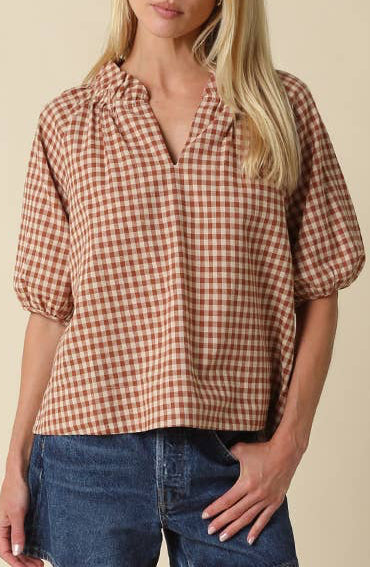A woman modeling a brown and beige gingham short-sleeve button-up shirt, paired with denim shorts and brown leather cowboy boots, against a solid beige background.