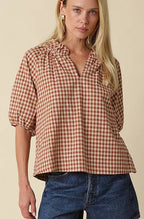 A woman modeling a brown and beige gingham short-sleeve blouse with a ruffled collar, paired with denim shorts and brown leather boots, against a solid beige studio background.