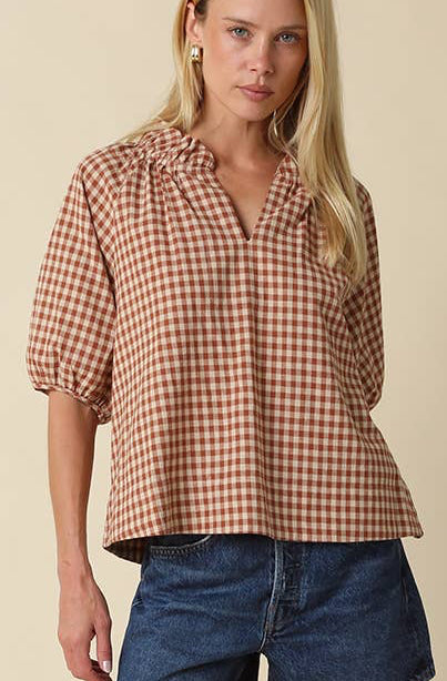 A woman modeling a brown and beige gingham short-sleeve blouse with a ruffled collar, paired with denim shorts and brown leather boots, against a solid beige studio background.