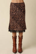 A leopard print midi skirt with a sheer overlay and black hem, styled with brown leather cowboy boots against a solid beige background.