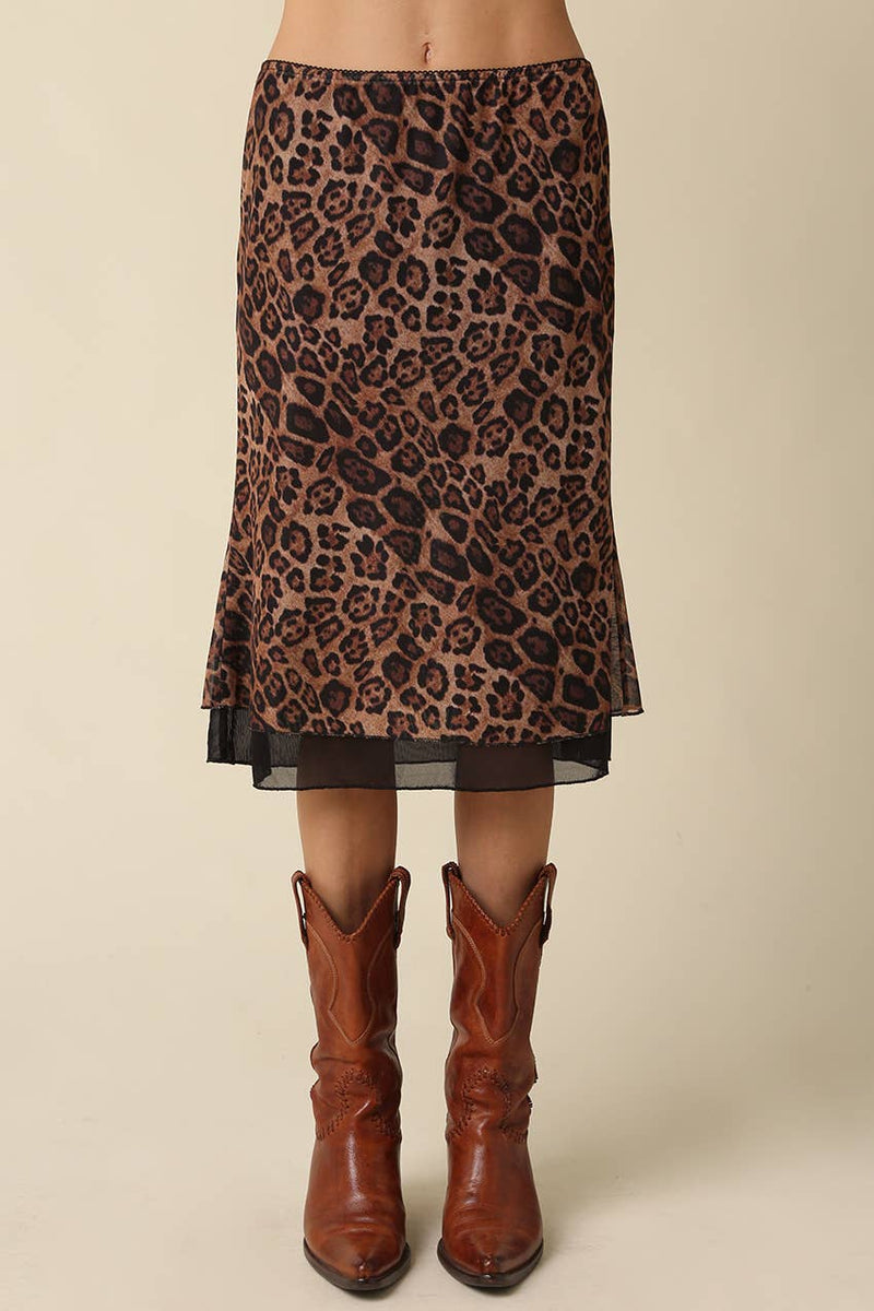 A leopard print midi skirt with a sheer overlay and black hem, styled with brown leather cowboy boots against a solid beige background.