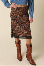 A woman modeling a blue cropped denim shirt and a brown leopard-print midi skirt, paired with brown leather cowboy boots, against a solid beige background.