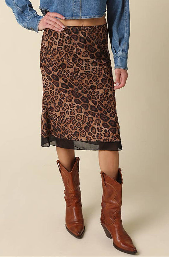 A woman modeling a blue cropped denim shirt and a brown leopard-print midi skirt, paired with brown leather cowboy boots, against a solid beige background.