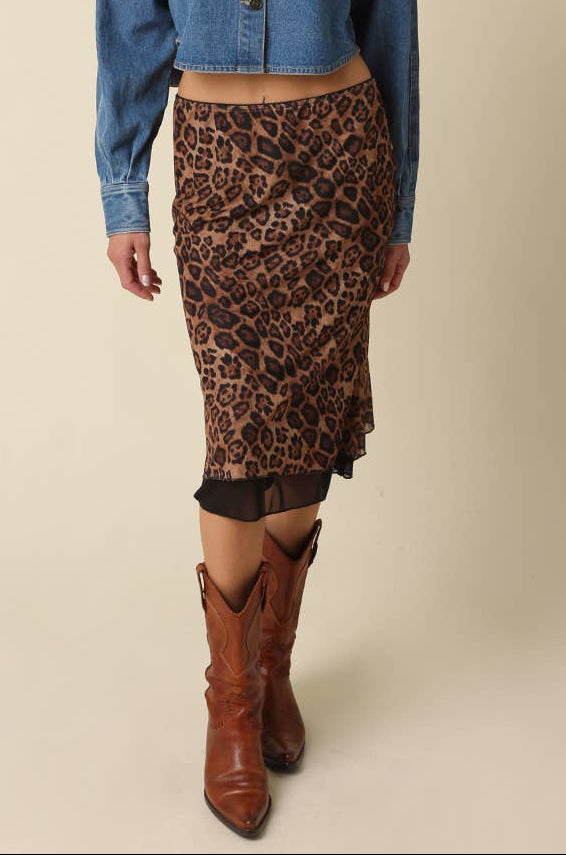 A woman modeling a blue cropped denim shirt-jacket and a brown leopard-print pencil skirt, paired with brown leather cowboy boots, against a solid beige studio background.
