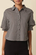 A woman with short curly black hair wearing a black and white vertically striped short-sleeve button-up shirt with puff sleeves, paired with black pants, against a solid beige background.