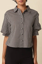 A woman with short curly black hair wearing a black and white vertically striped short-sleeve button-up shirt with puff sleeves, paired with black pants, against a solid beige background.
