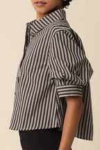 A woman with short curly black hair models a black and white vertically striped short-sleeve button-up shirt, paired with black pants, against a solid beige background.